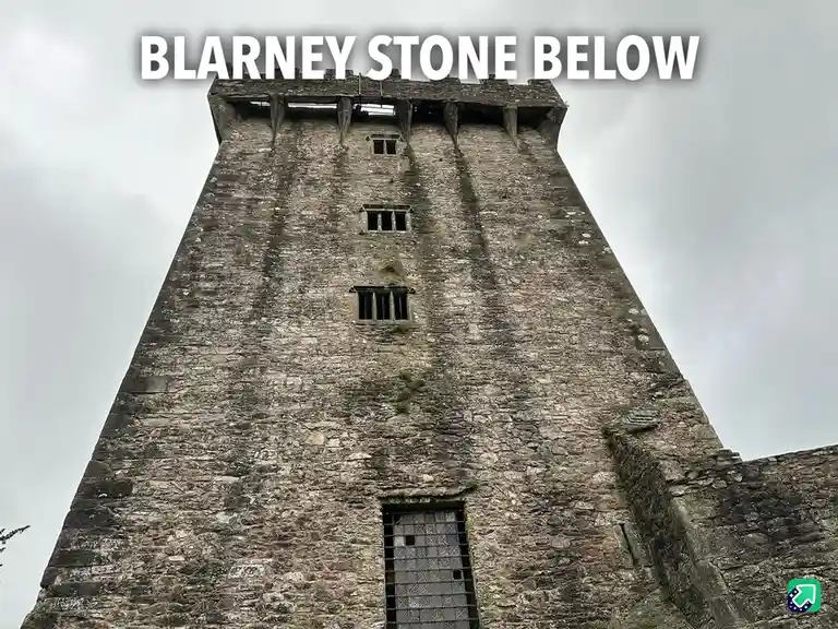 A low-angle photo shows the tall, weathered stone tower of Blarney Castle against a cloudy sky. The image has large white…