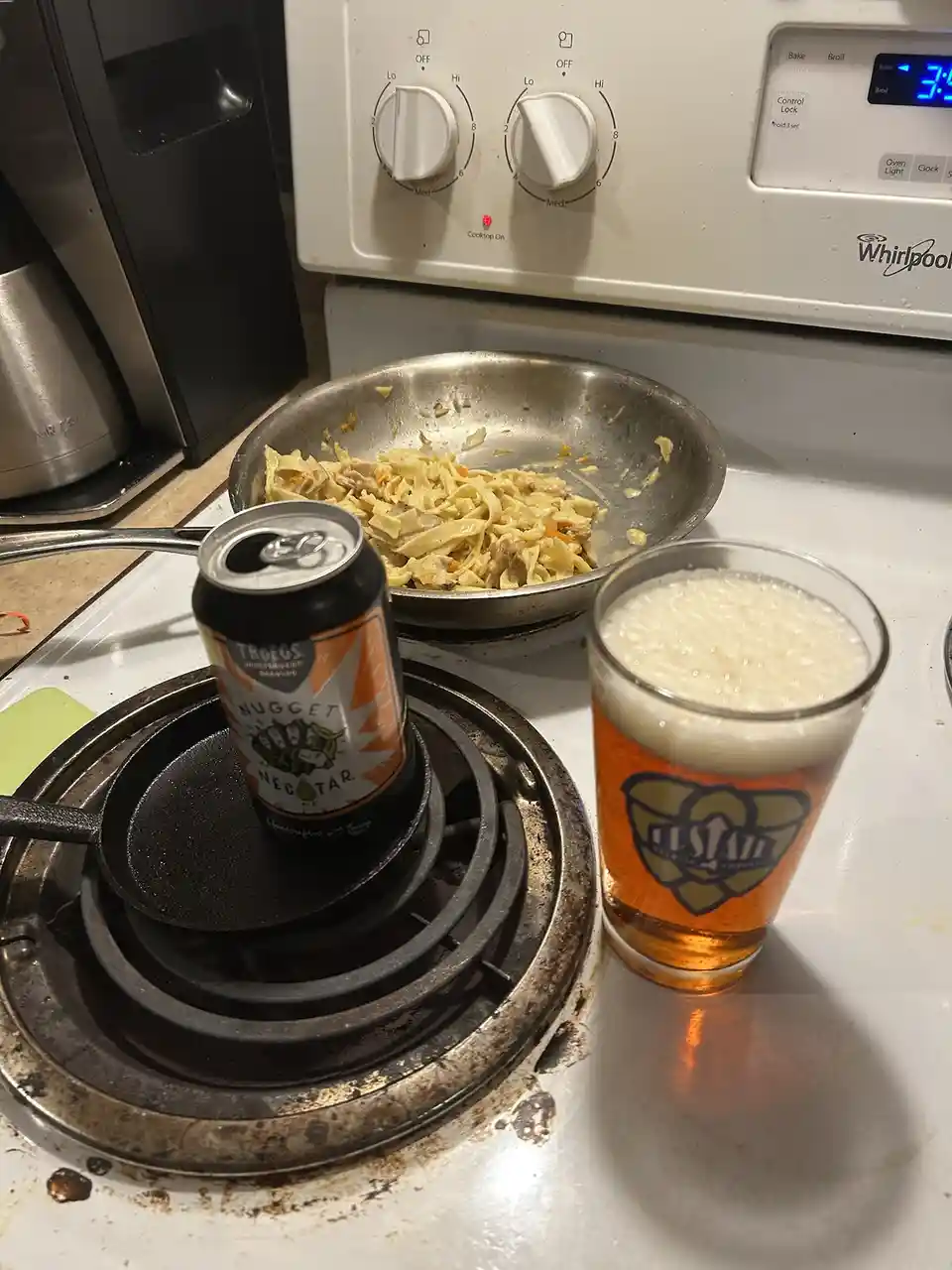 A photo of a kitchen stove with a nearly empty frying pan of pasta, a can of Troegs Nugget Nectar Ale sitting on a cast iron…