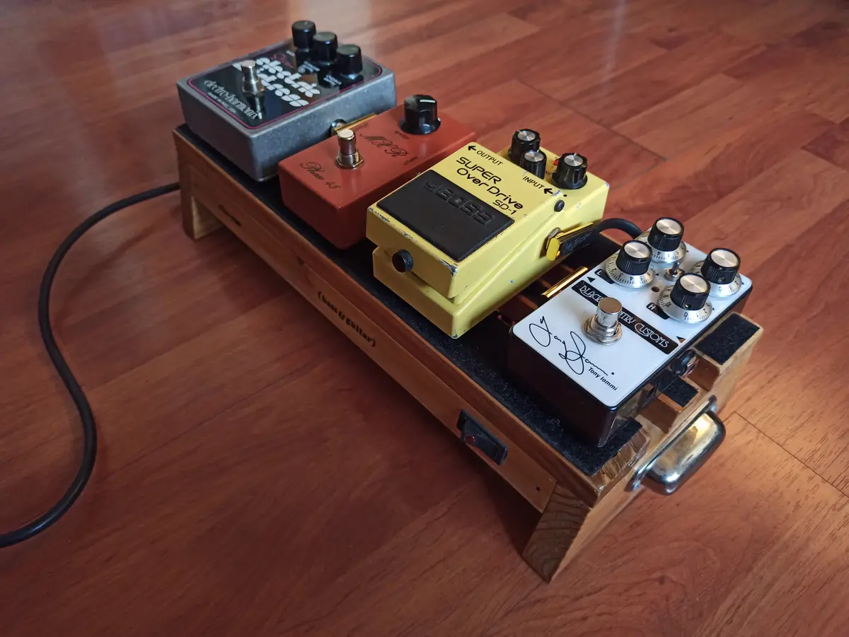 A wooden pedalboard with four guitar effects pedals — a grey Electro-Harmonix, a red Phase 95, a yellow Boss SD-1 Super…