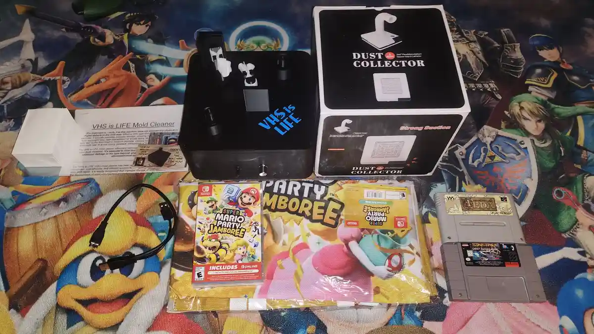 A flat lay of retro gaming and VHS care items on a colorful anime-themed blanket, including a 'VHS is LIFE' mold cleaner…