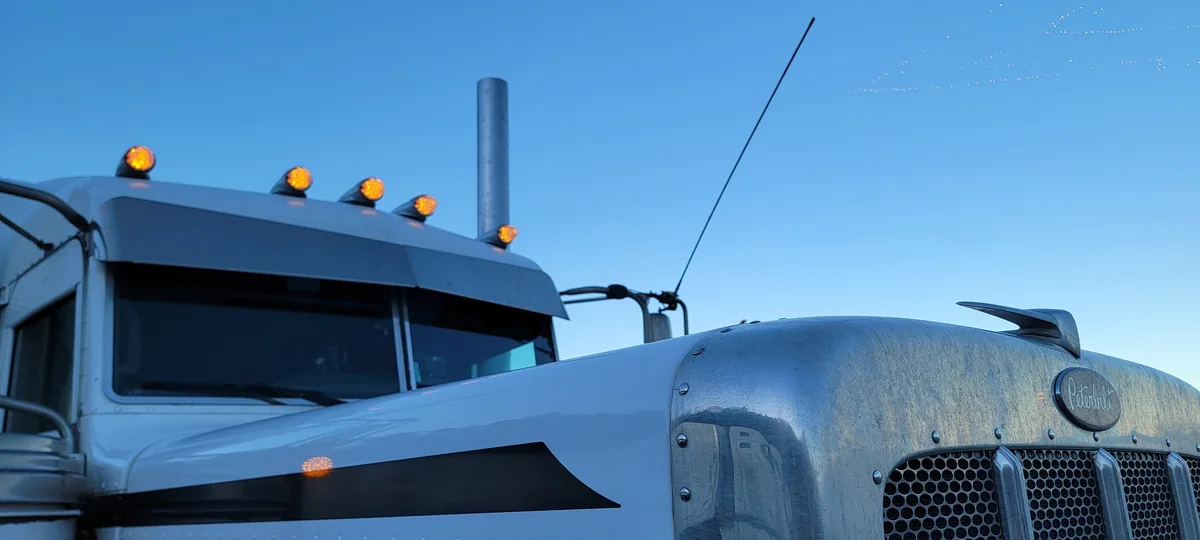 A low-angle photo of a silver Peterbilt semi-truck cab against a clear blue sky, with its roof-mounted amber lights glowing…