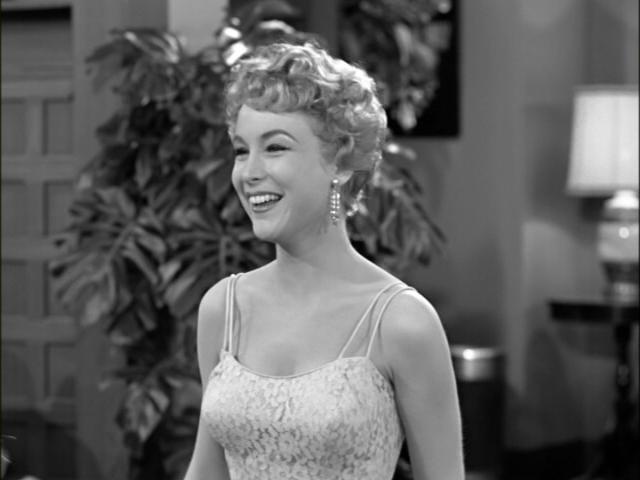 a black-and-white still of a smiling woman with curly hair wearing a lace dress and dangling earrings, overlaid with the…