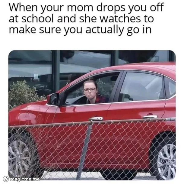 A woman with glasses sits in the driver's seat of a red car parked behind a chain-link fence, looking intently forward with…