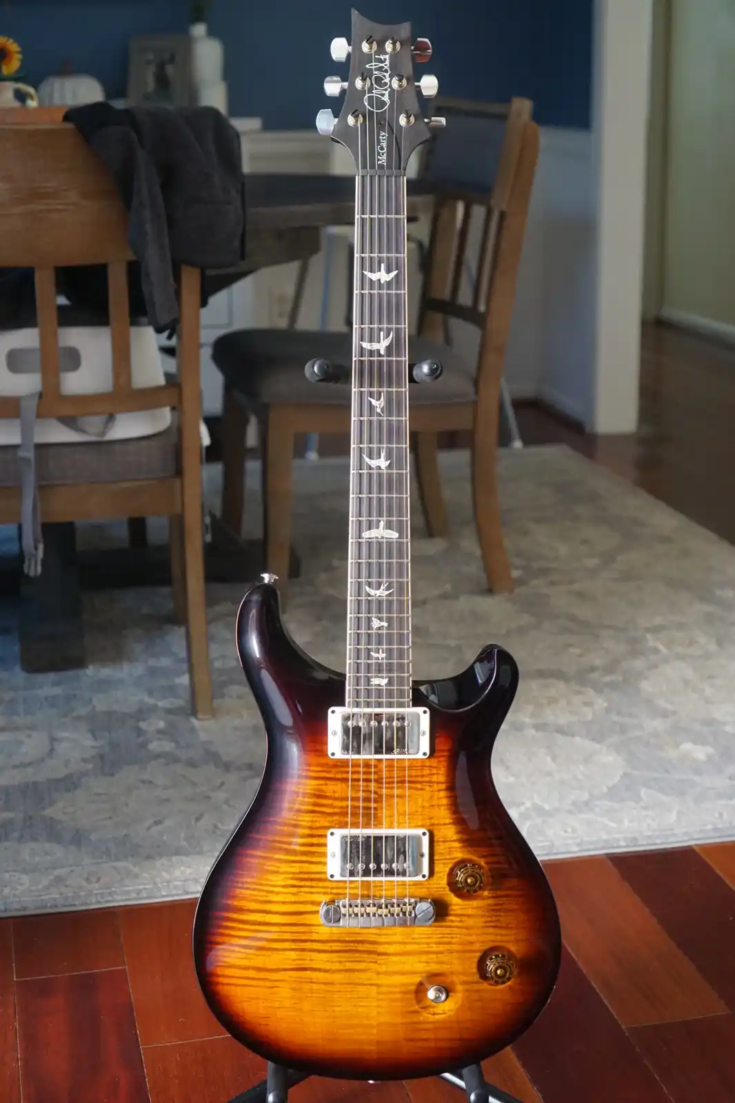 A sunburst-finish electric guitar with a bird in flight inlay on the fretboard stands upright on a stand in a home setting…