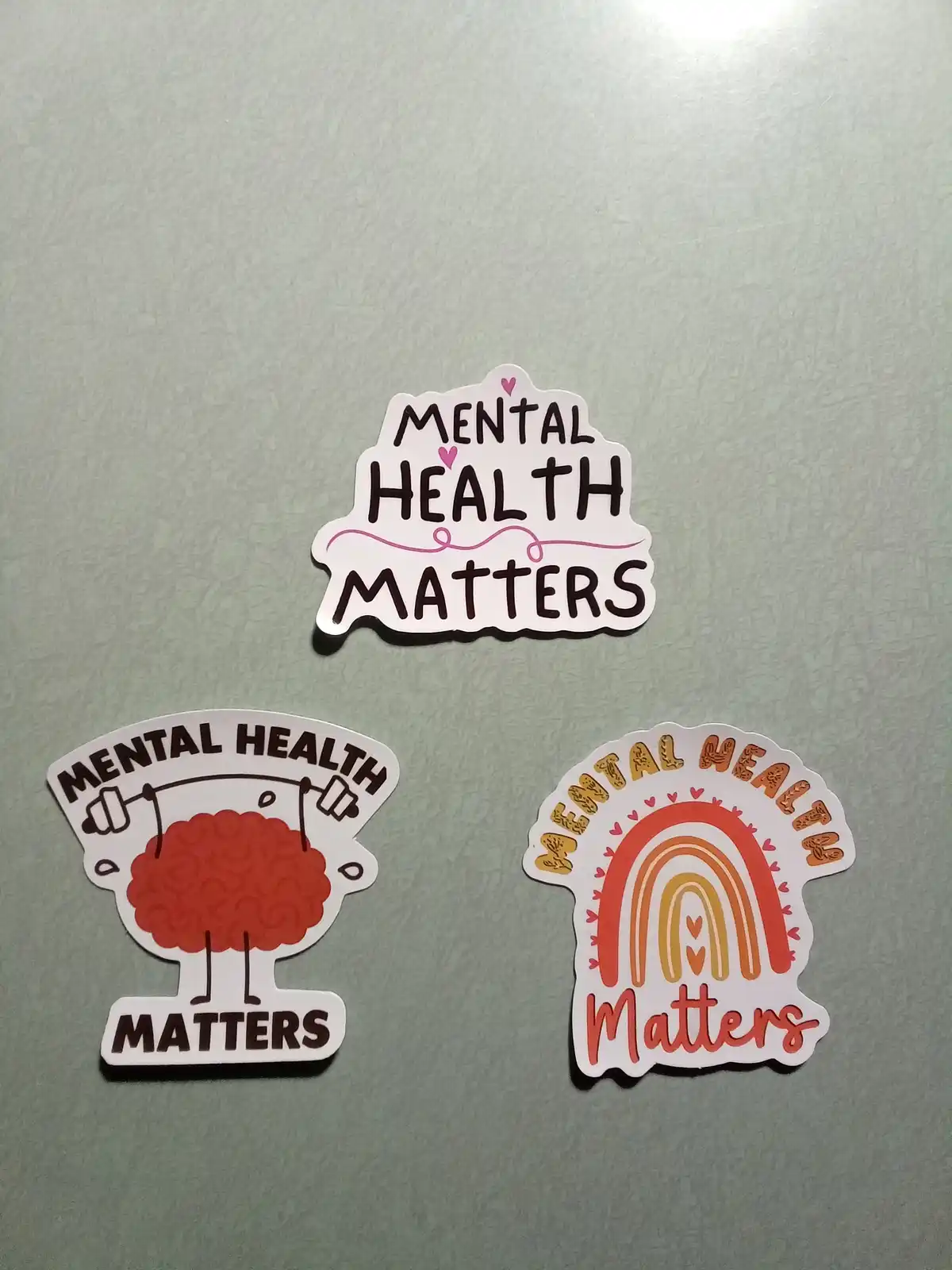 Three 'Mental Health Matters' stickers are arranged on a light gray surface. The top sticker has bold black text with pink…