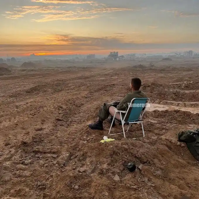 A man in military-style clothing sits in a blue folding chair on a barren, muddy field at sunset, facing a distant, hazy…