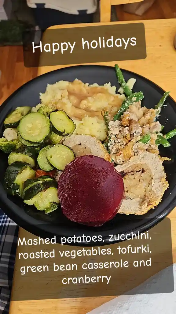 A black plate holds a holiday meal featuring mashed potatoes with gravy, sliced roasted zucchini, a large round beet, sliced…