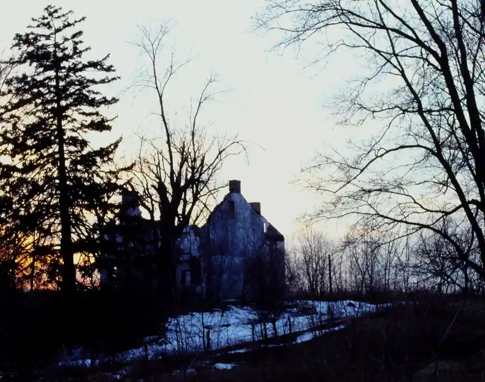 A dilapidated, snow-dusted house stands in silhouette against a twilight sky, framed by bare trees and a distant orange…