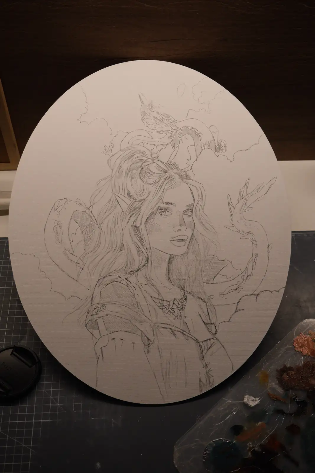 A pencil sketch on an oval canvas depicts a fantasy woman with long hair and pointed ears, surrounded by dragons in the sky…