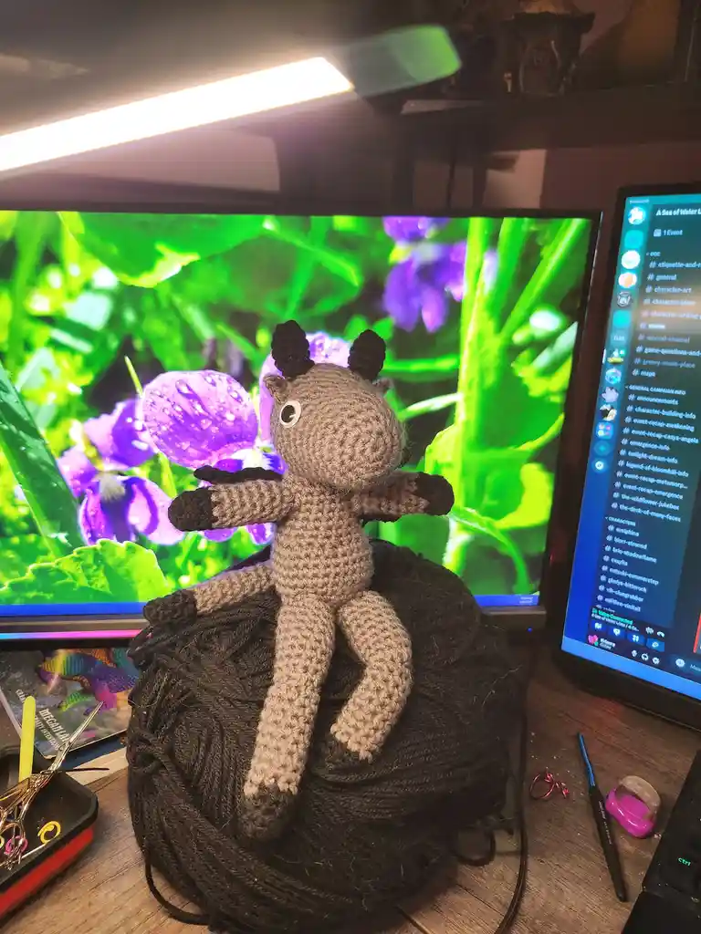 A handmade crocheted giraffe plushie sits atop a large ball of dark yarn on a wooden desk, with a computer monitor…