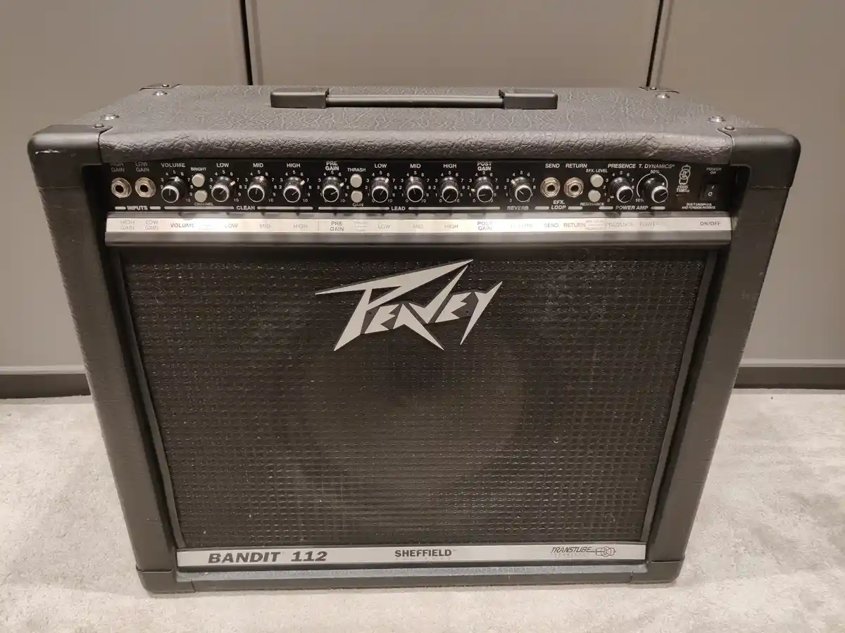A black Peavey Bandit 112 guitar amplifier sits on a light-colored carpet, featuring a detailed control panel with numerous…