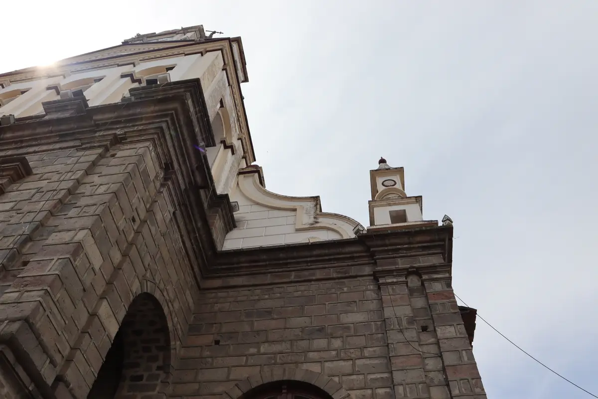 A low-angle photo shows the corner of a stone church building with a bell tower against an overcast sky. The image is…