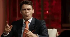 A man in a dark suit and orange tie sits in a white chair against a red background, his expression shifting from intense to…