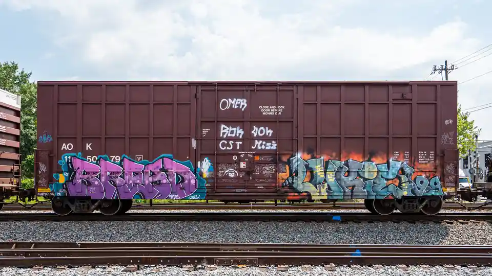 A brown freight train car covered in colorful graffiti, including large purple and blue tags, sits on railway tracks under a…