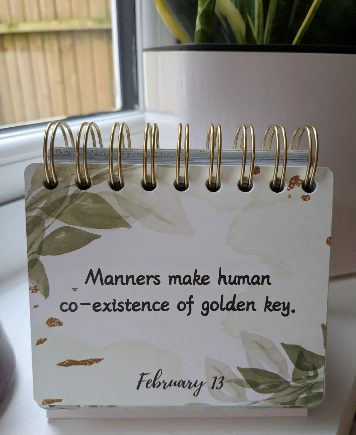 A close-up of a spiral-bound desk calendar with a leafy watercolor design and gold glitter accents, sitting on a windowsill…