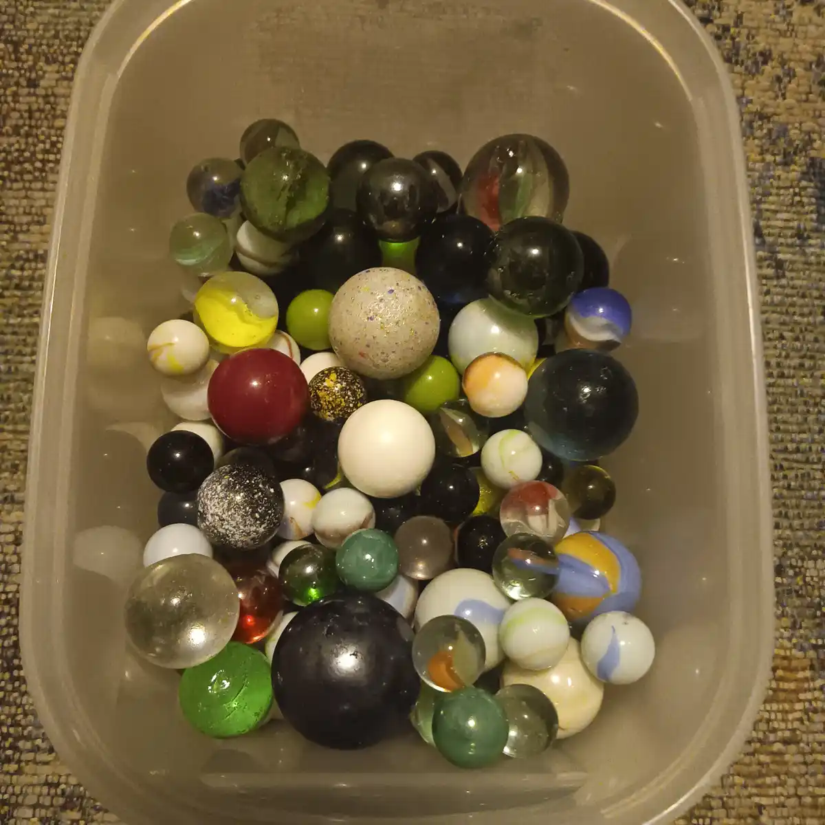 A top-down view of a clear plastic container filled with a large, colorful assortment of marbles of various sizes, patterns…