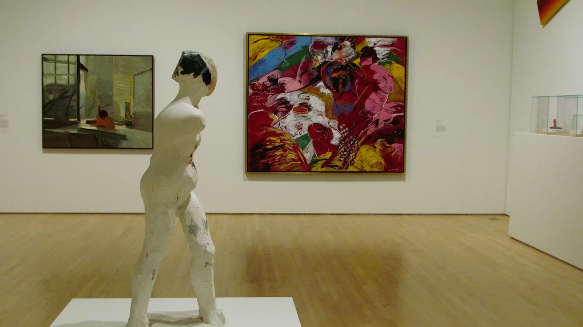 A white, unfinished-looking sculpture of a human figure stands in an art gallery, facing a large, colorful, abstract…