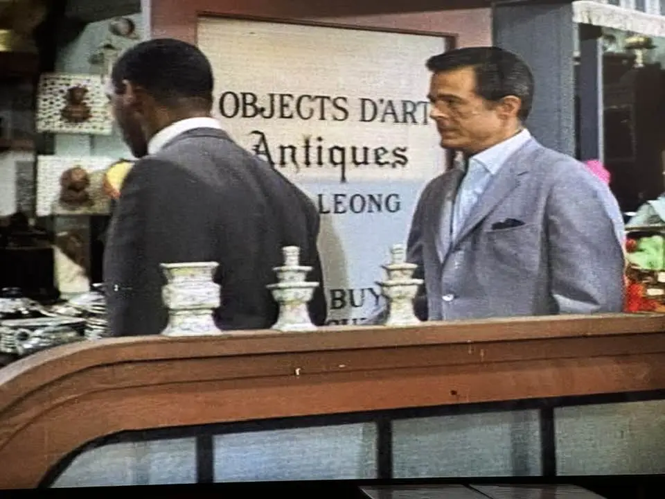 Two men stand behind a counter in an antique shop with a sign reading 'OBJECTS D'ART Antiques LEONG' visible behind them…