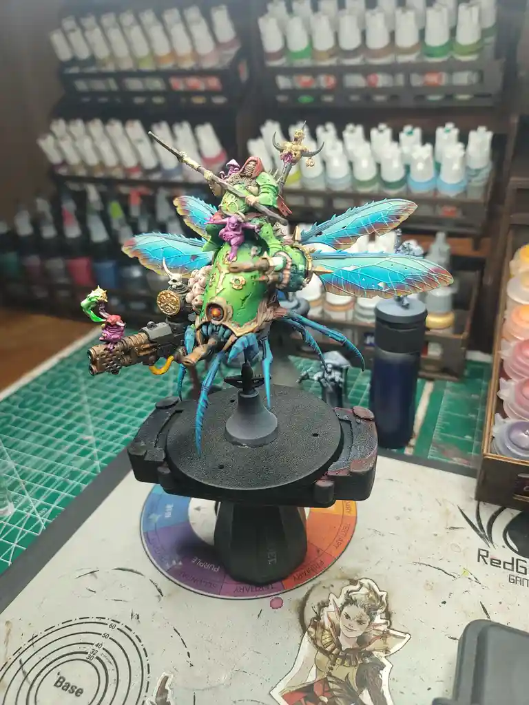 A highly detailed, painted miniature of a green, insectoid warlord with large blue wings and multiple weapons sits on a…