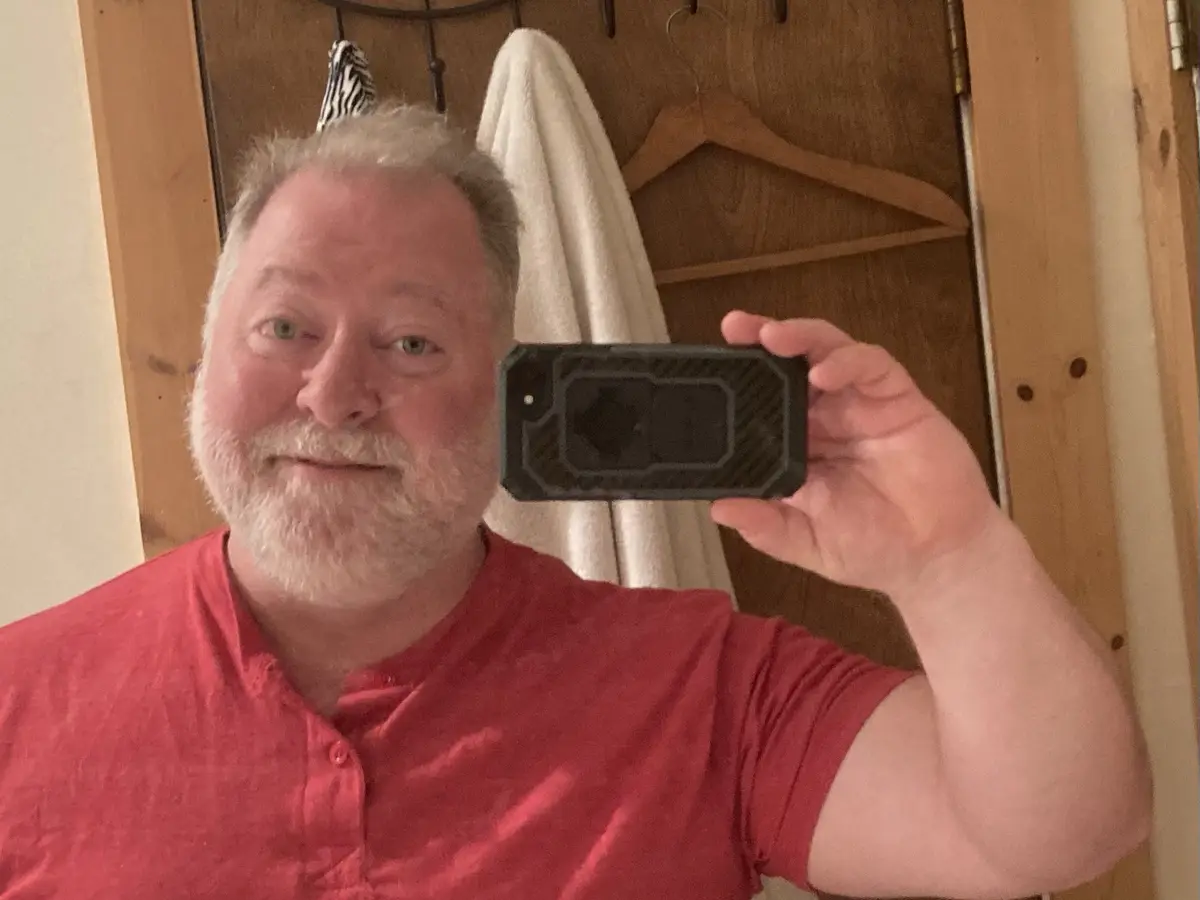 A middle-aged man with a white beard and red shirt takes a selfie in a mirror, holding up his phone with a black rugged…
