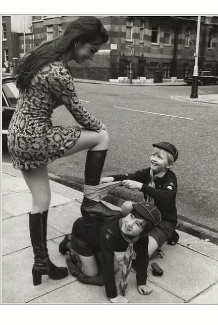 A black-and-white photo shows a woman in a patterned mini dress and tall boots standing on a sidewalk, smiling down as two…