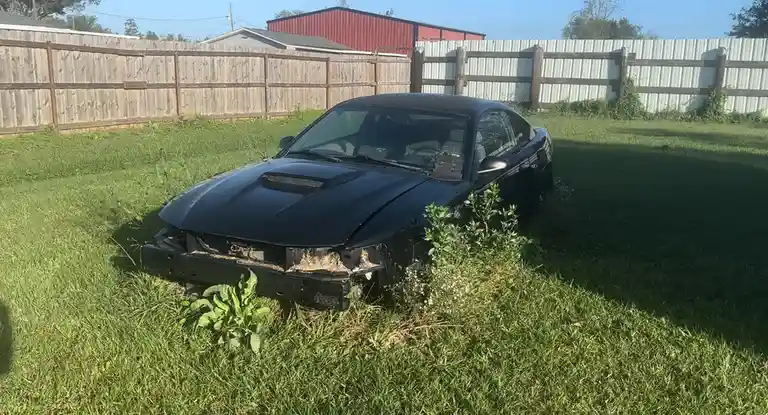 A black sports car with a severely damaged front bumper and missing grille sits abandoned in a grassy yard, partially…
