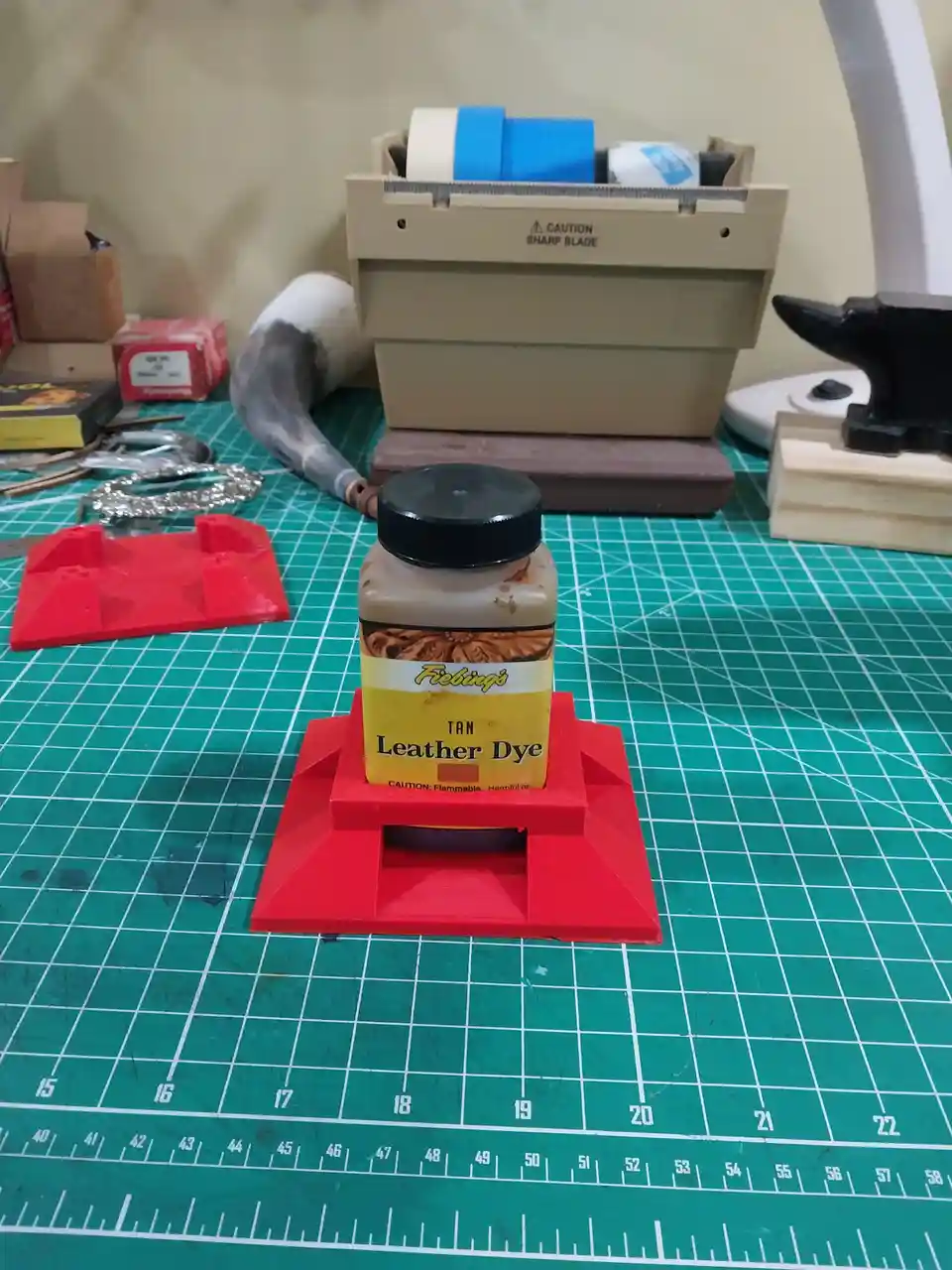 A jar of Fiebing's Tan Leather Dye sits on a red 3D-printed stand on a green cutting mat, surrounded by crafting tools like…
