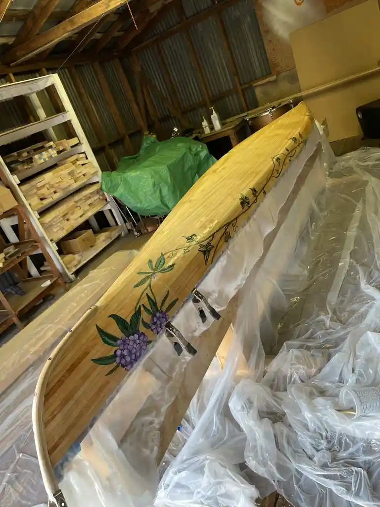 A wooden canoe with hand-painted purple flowers and green vines is propped up in a workshop, partially covered in plastic…
