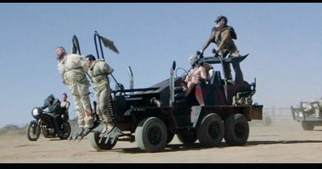 A scene from Mad Max: Fury Road shows a heavily modified, six-wheeled vehicle with several armed, rugged individuals. Two…