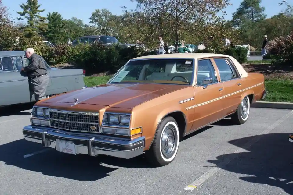 A brown and beige 1970s Buick Regal sedan is parked in a lot with people around. The overlaid text reads 'i'm not a car guy…