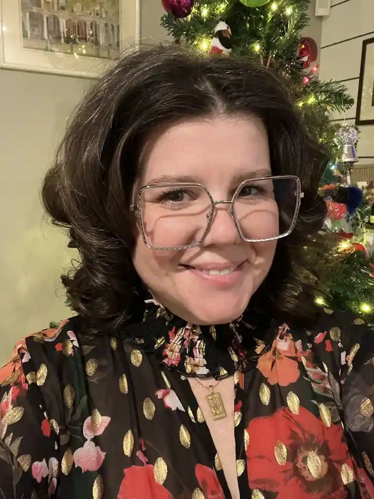 A woman with dark curly hair and large square glasses smiles at the camera while wearing a black floral and gold-patterned…