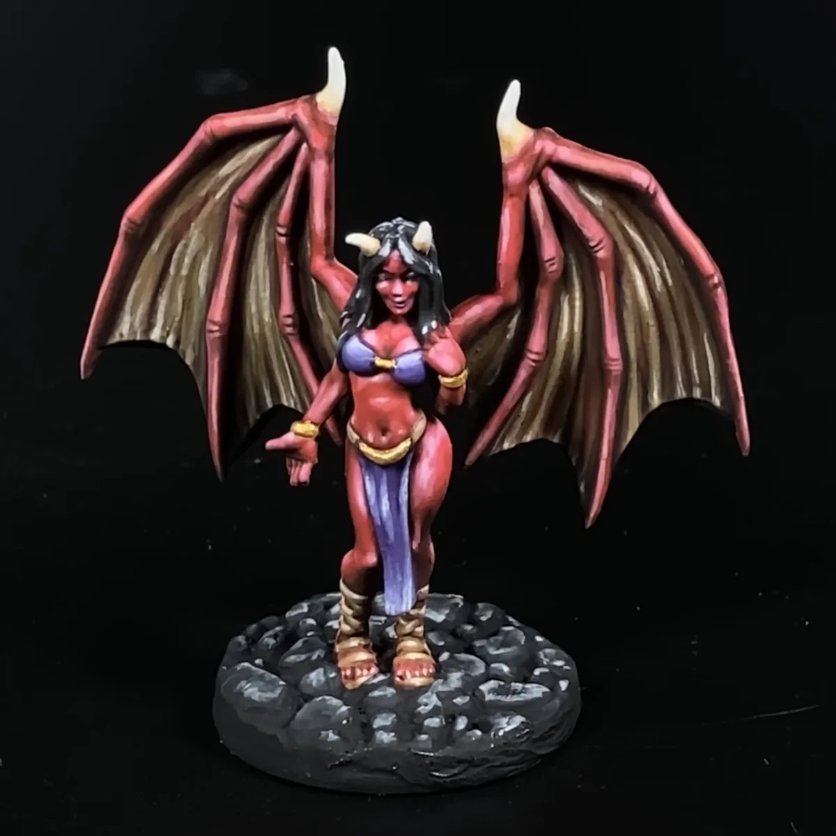 A painted miniature figurine of a female demon with red skin, black hair, and small horns, standing on a rocky base with…