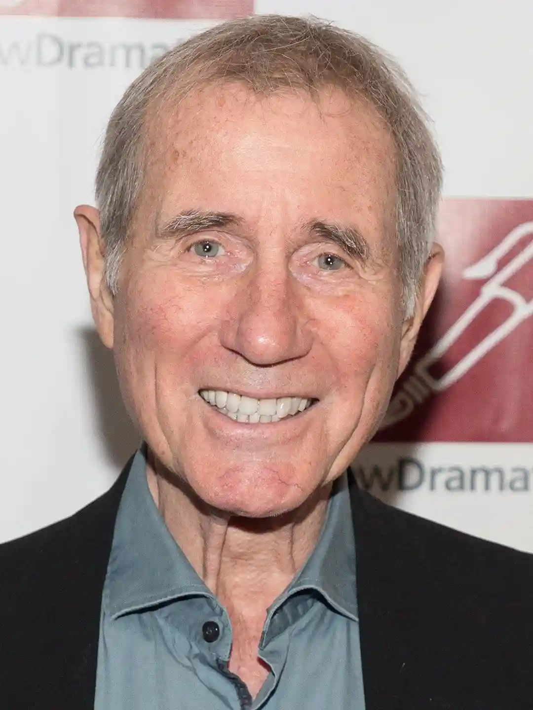 A close-up photo of an older man with gray hair, wearing a dark jacket over a light blue collared shirt, smiling broadly at…