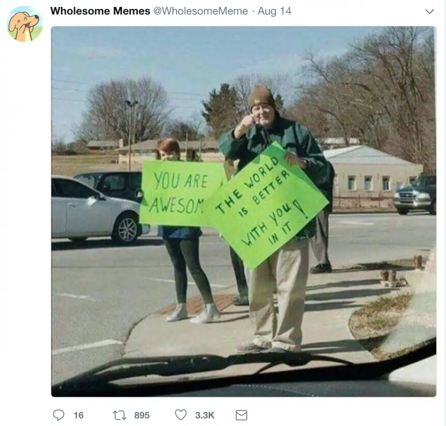 Two people stand on a sidewalk holding bright green signs. The person in front holds a sign that reads 'YOU ARE AWESOME.'…
