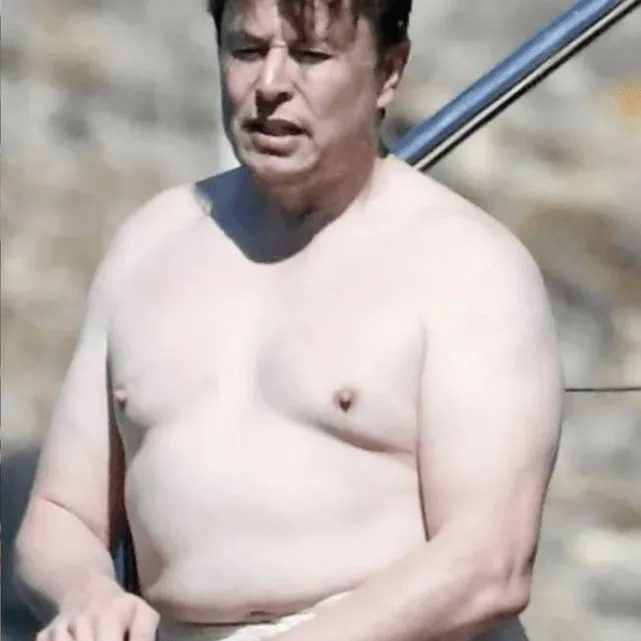 A shirtless man with a round belly and a tired expression stands outdoors, partially shaded, with a metal railing visible…