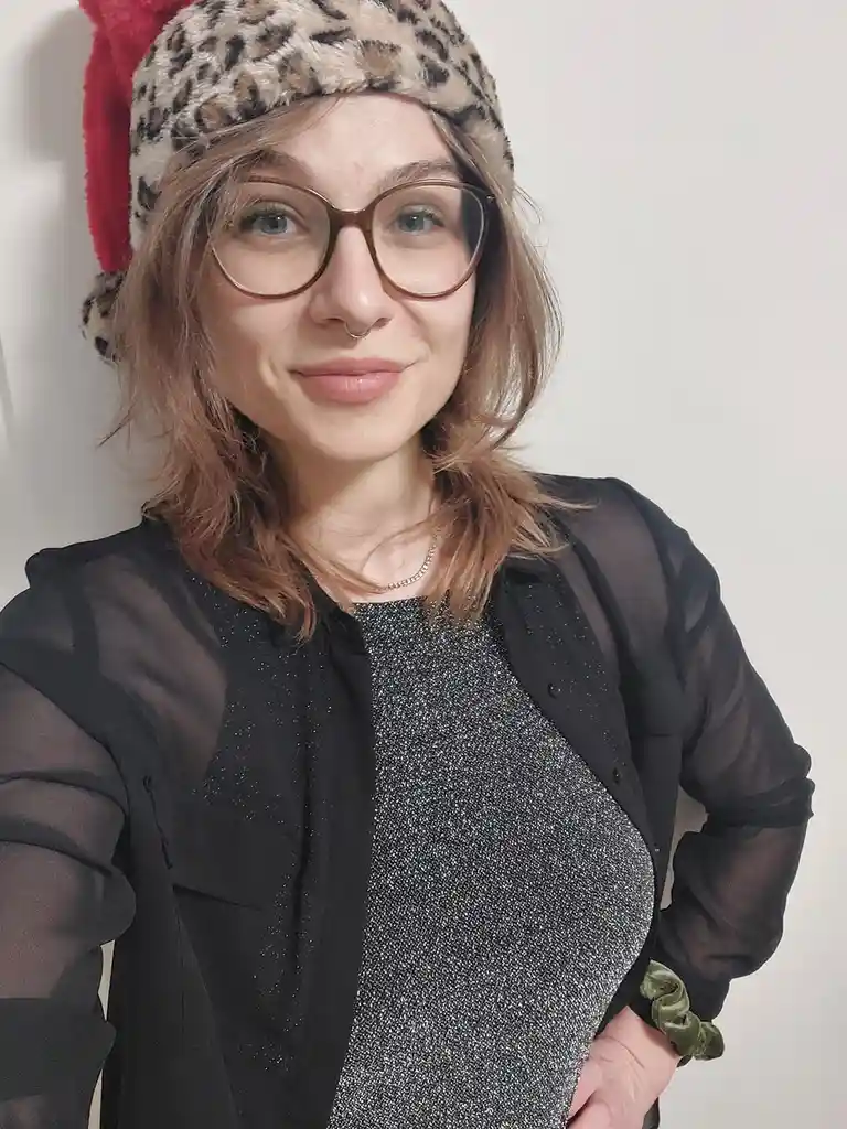 A woman with glasses and a nose ring wears a Santa hat with a leopard print pattern and a red pom-pom, smiling at the…