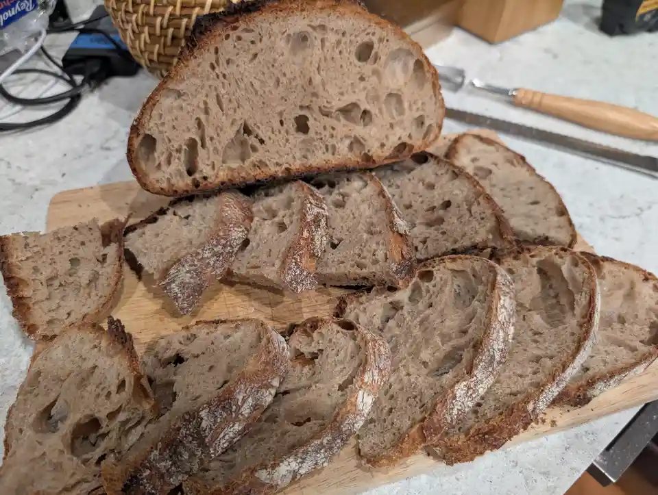 A wooden cutting board holds numerous thick slices of rustic, dark brown sourdough bread with a visibly porous crumb and…