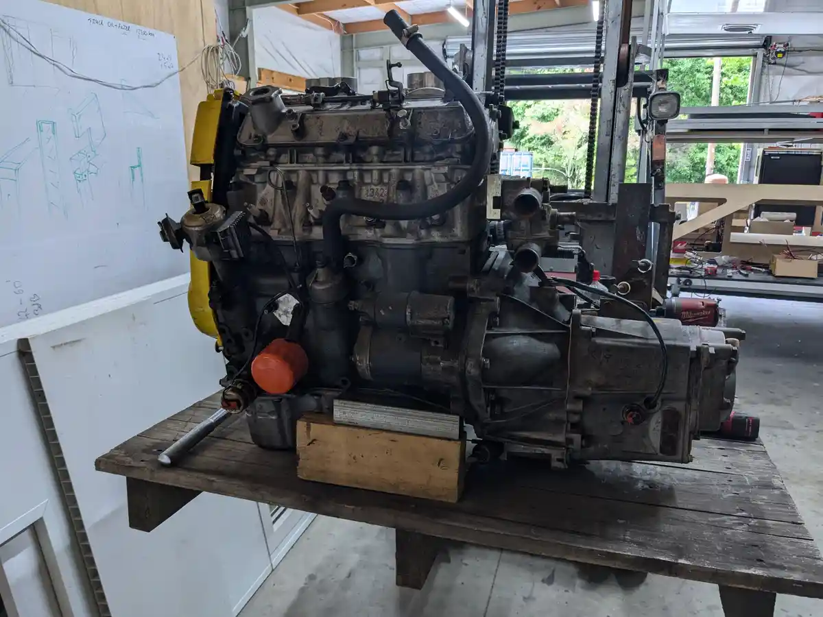 A large, dirty, disassembled truck engine with a transmission attached sits on a wooden workbench in a cluttered garage. The…