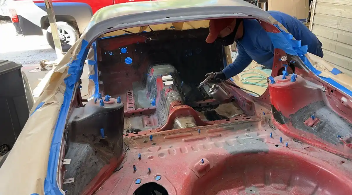 A person wearing a blue shirt, red cap, and face mask is spray painting the interior of a car's body panel. The car's…