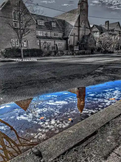 A black-and-white photo of a brick church and street, with a vibrant blue puddle in the foreground reflecting the sky and…