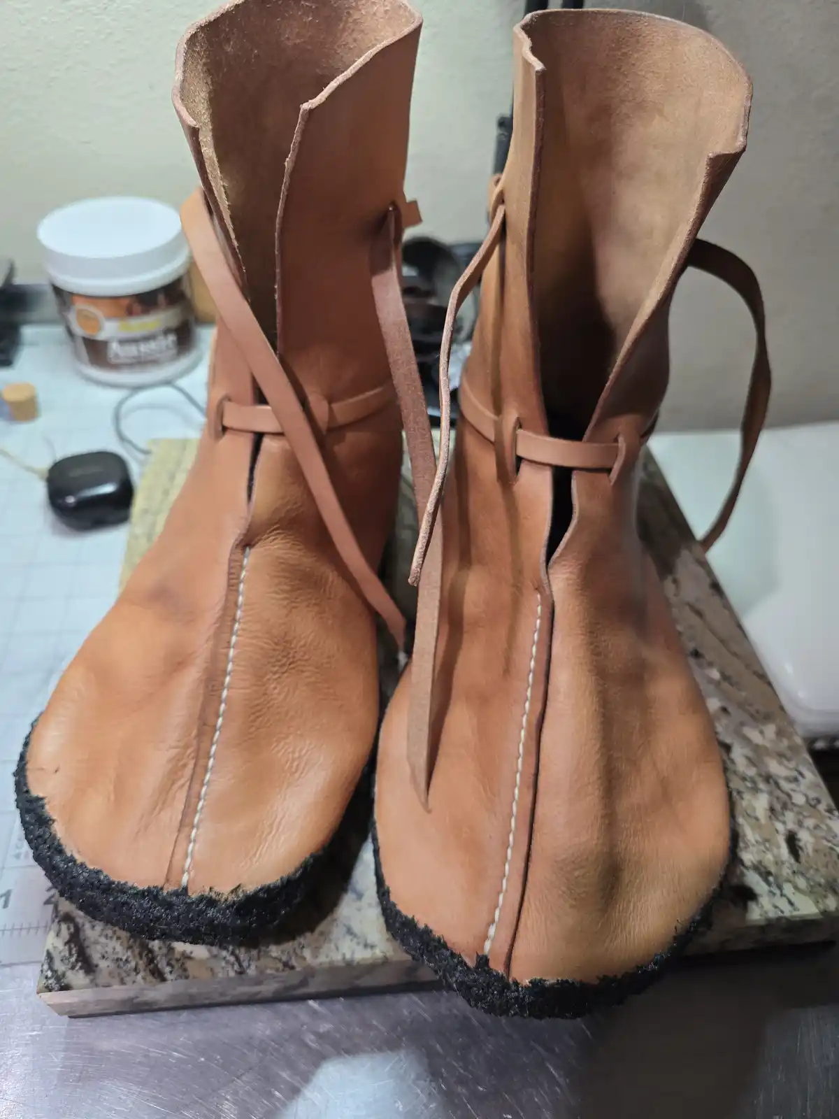 A pair of handmade tan leather boots with laces and white stitching, sitting on a granite surface. The boots have a raw…