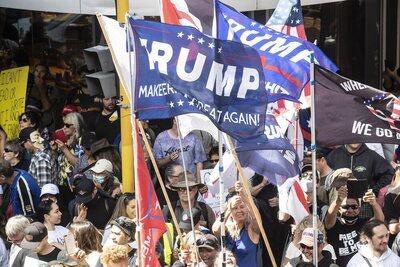 A crowd of people at a political rally holds multiple blue and red flags. One large blue flag prominently displays 'TRUMP…