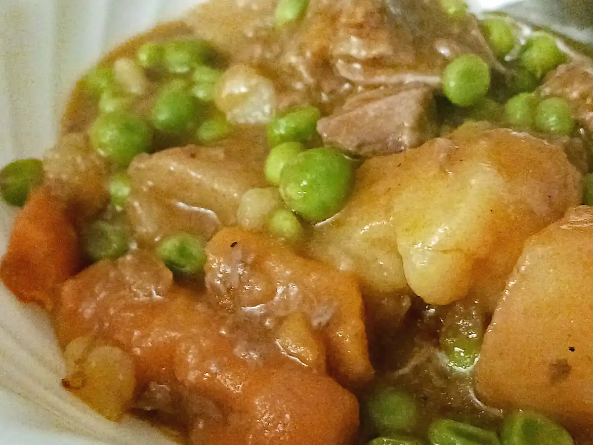 a close-up photo of a hearty stew in a white bowl, featuring chunks of meat, potatoes, carrots, and bright green peas…
