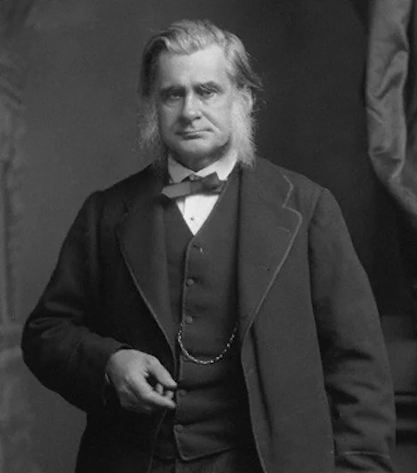 A black-and-white portrait of a stern-looking older man with long hair, wearing a formal suit, vest, bow tie, and chain…