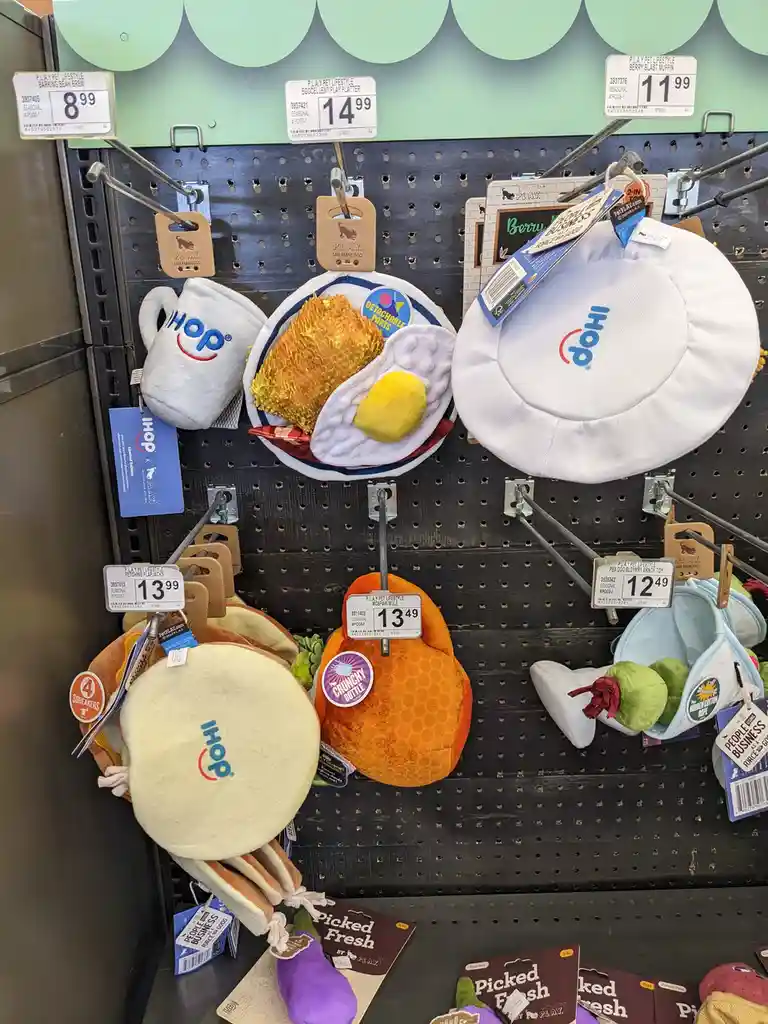 A store shelf displays various IHOP-themed pet toys shaped like breakfast foods such as a mug, an egg dish, a pancake, a…