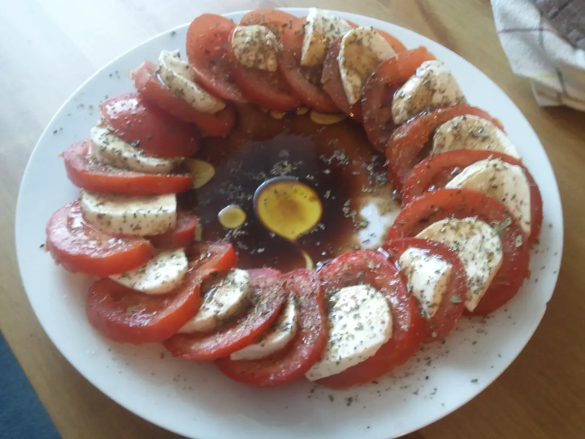 A white plate holds a circular arrangement of sliced red tomatoes and white mozzarella cheese, drizzled with dark balsamic…