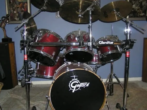 A full drum kit with red and black drums, chrome hardware, and cymbals is shown in a room. The bass drum has a 'Gretsch'…
