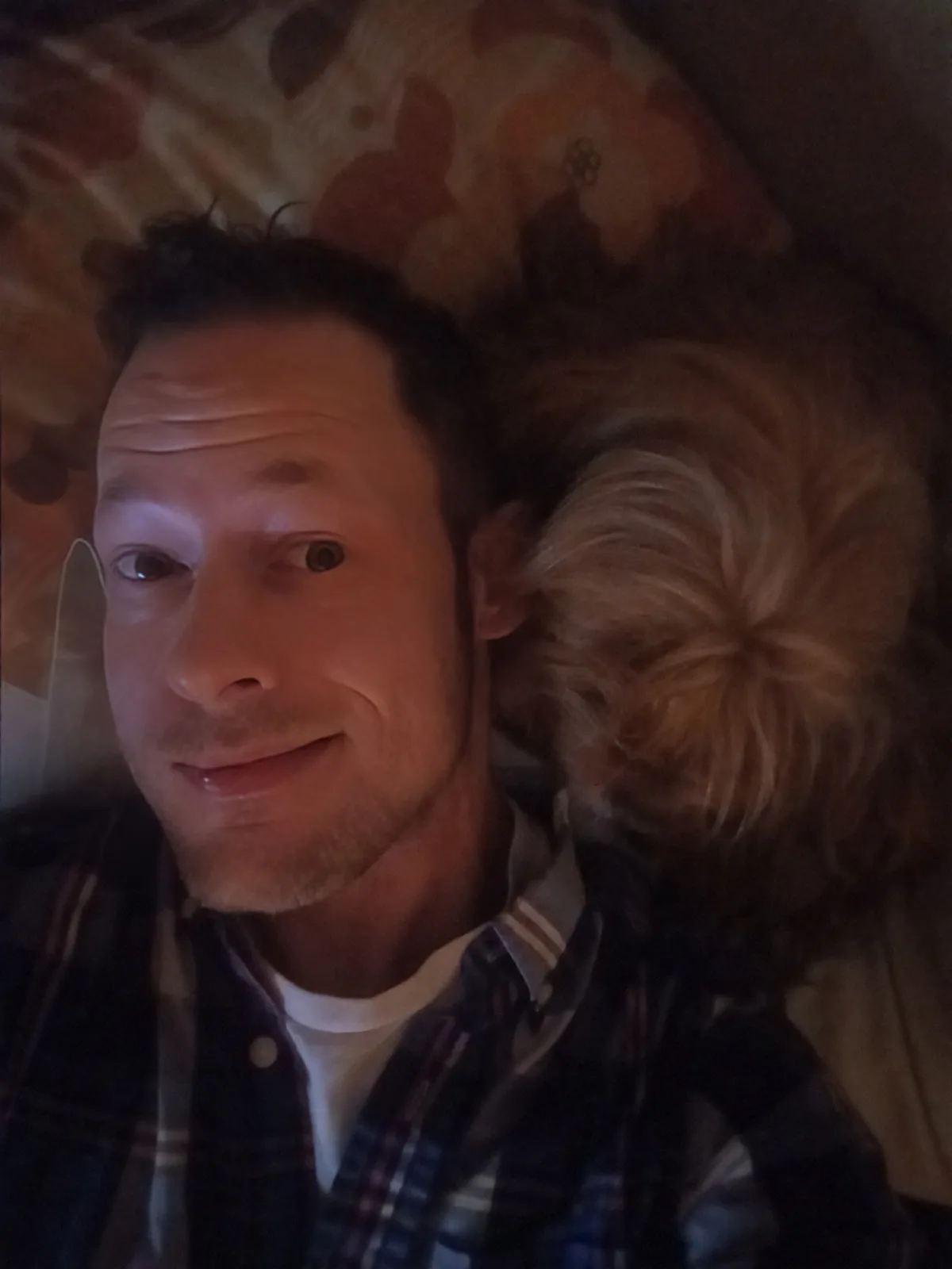 A man with short dark hair and a slight smile lies on his back on a floral-patterned couch or bed, wearing a plaid shirt…