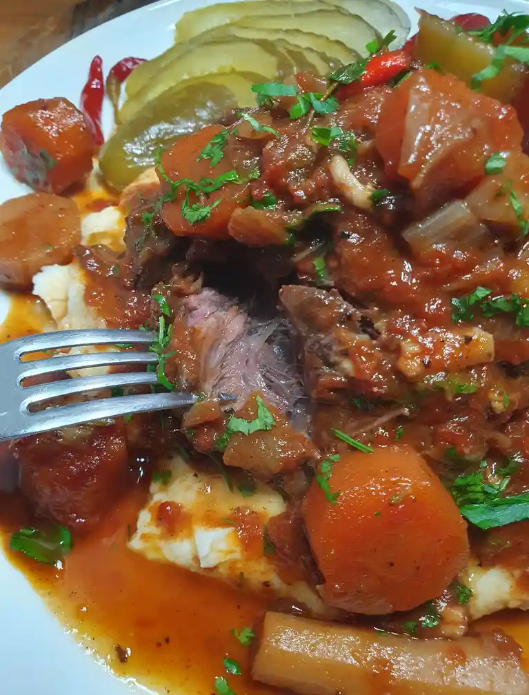 A close-up photo of a plate of stew with shredded meat, carrots, pickles, and mashed potatoes, with a fork piercing the…