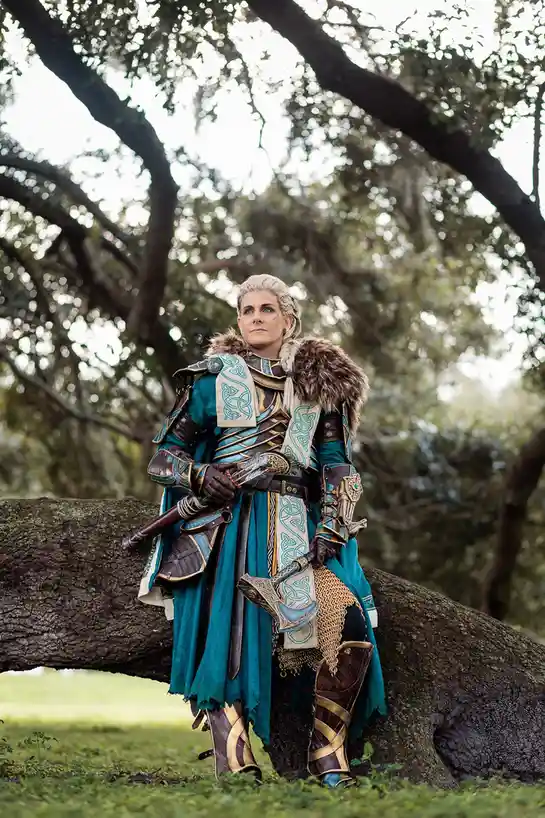 A person in elaborate fantasy warrior cosplay, featuring teal and brown armor with fur trim, chainmail, and a sword, stands…